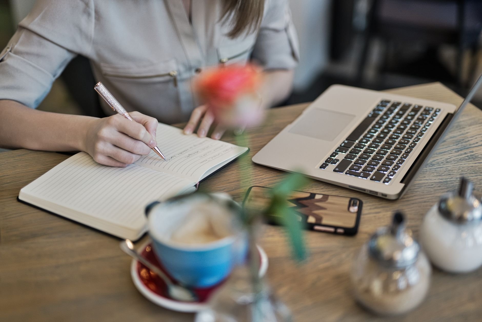 Woman writing in a notebook with a laptop and coffee on a desk — the daily work of building a newsletter